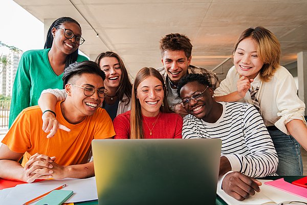 Studierende befinden sich entweder sitzend oder stehend an einem Tisch und schauen gemeinsam auf einen Laptop. Die Kamera ist frontal auf sie gerichtet-