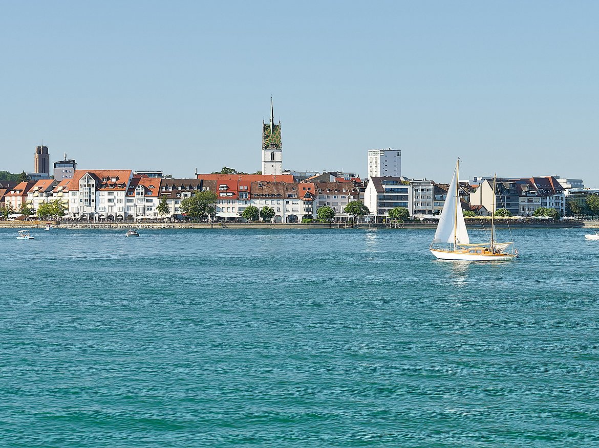 Anblick der Stadt Friedrichshafen am Bodensee. Vorne sieht man den Bodensee, im Hintergrund befindet sich Friedrichshafen.