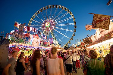 Zu sehen ist eine Menschenmasse auf einem Jahrmarkt. Im Hintergrund befinden sich beleuchtete "Buden" und ein Riesenrad.