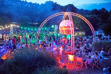 ZU sehen sind viele Personen in einem Park. Zentral befindet sich eine offene Kuppel mit Discokugel. Alles leuchtet bunt.