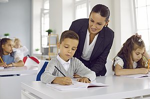 Kinder in Schulklasse; eine Lehrerin beugt sich zu einem Jungen, der schreibt.