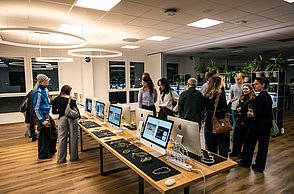 Menschengruppe steht vor einem Tisch auf dem viele Computer stehen. Auf den Bildschirmen werden Bilder gezeigt.
