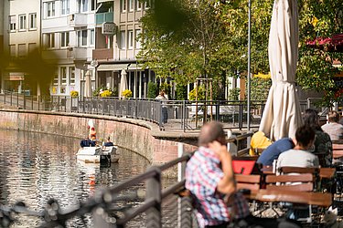 Auf der linken Seite sieht man die Enz, auf der sich ein Tretboot mit Personen befindet. Auf der rechten Seite sitzen Personen an Tischen.